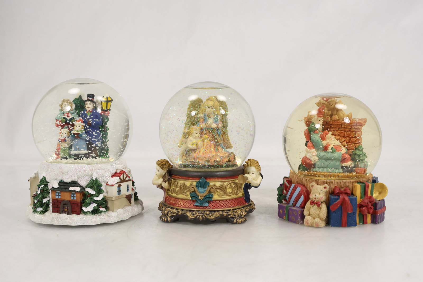 Collection of Three Christmas Snow Globes with Holiday Figures: Group of three decorative Christmas snow globes featuring festive holiday scenes beneath clear domes. Included are a winter snowman scene, an angelic or nativity-inspired holiday figure group, and a C