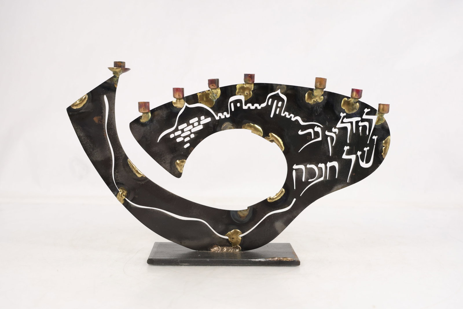 Handcrafted Jerusalem-Themed Metal Hanukkah Menorah with Hebrew Inscription: Handcrafted metal Hanukkah menorah featuring a contemporary sculptural form accented with a stylized skyline of Jerusalem and Hebrew lettering. The curved body supports nine candle cups, including the