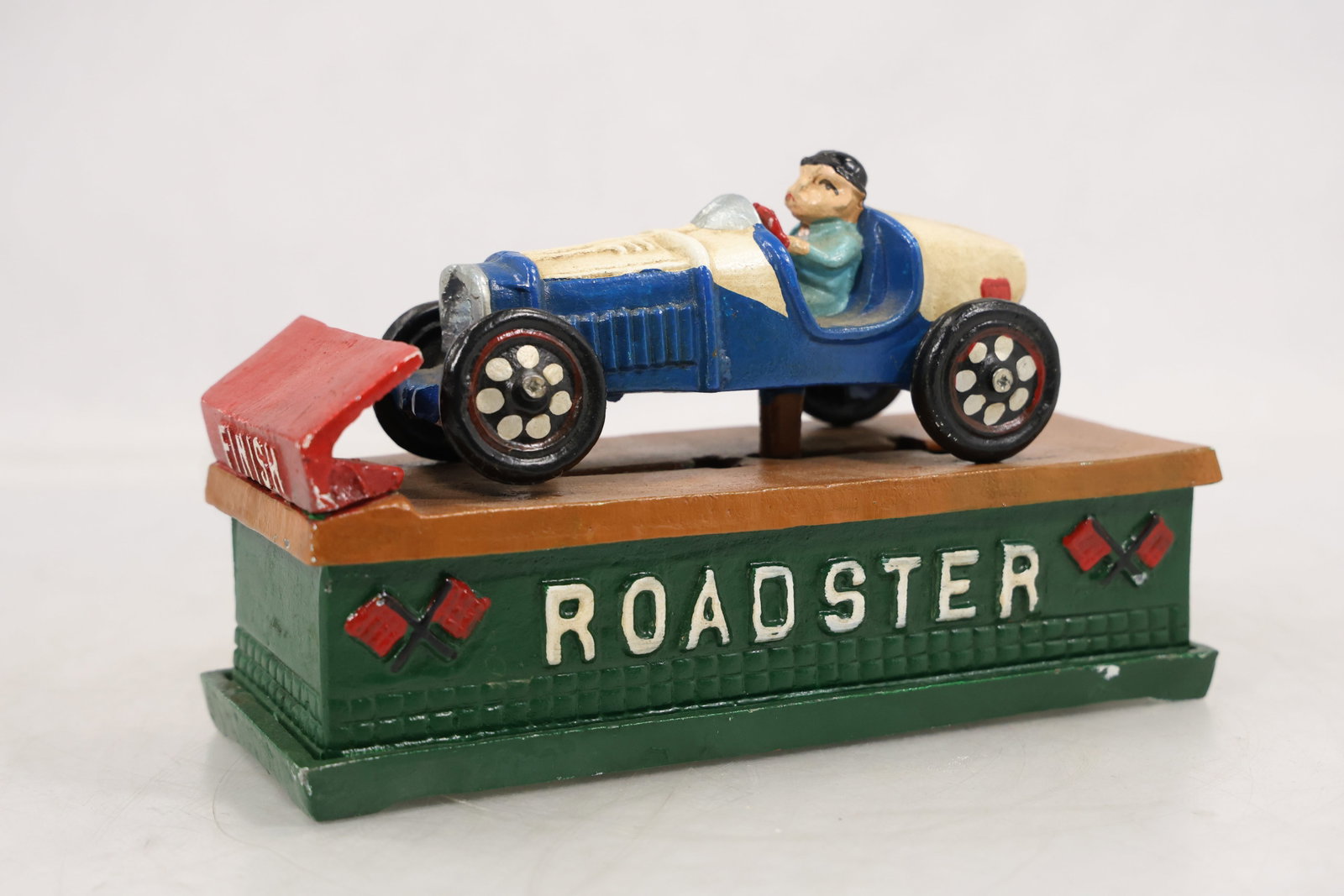 Iron Mechanical Bank Roadster Racing Car with Driver and Finish Gate: ainted cast iron mechanical bank in the form of an early racing roadster mounted on a rectangular base with raised lettering reading Roadster. The car is finished in blue and cream tones with a seated