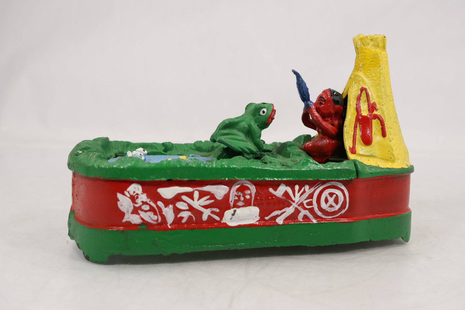 Cast Iron Mechanical Coin Bank Teepee and Frog Scene with Painted Figures: Cast iron mechanical coin bank depicting a scene with a teepee structure, a seated figure holding an object, and a frog positioned on a textured ground surface. The base is elongated and painted green