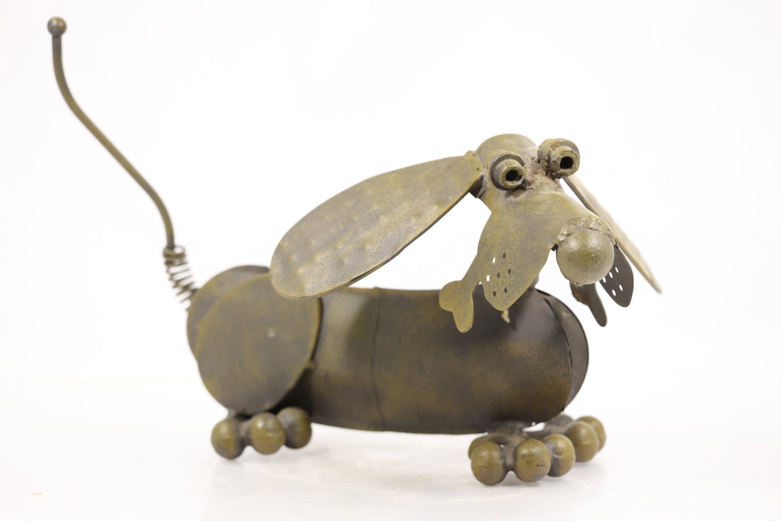 Whimsical Metal Dachshund Dog Sculpture: A whimsical elongated dachshund dog is assembled from metal components with a long low body short legs curled tail and modeled head. The dark patinated surface and playful proportions are visible in t