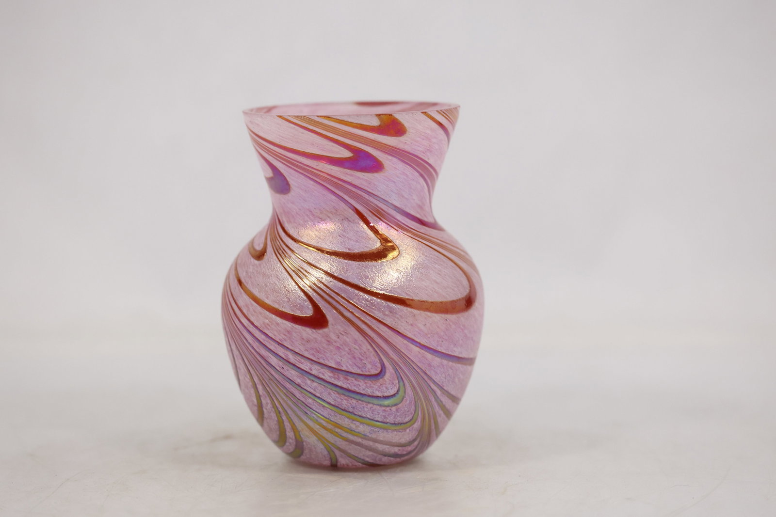 Teleflora Iridescent Pink Swirl Art Glass Vase: A Teleflora iridescent art glass vase has a rounded baluster body a narrowed neck and a flared rim. Frosted pink glass is wrapped with pulled feather ribbons in purple white green and gold tone colors