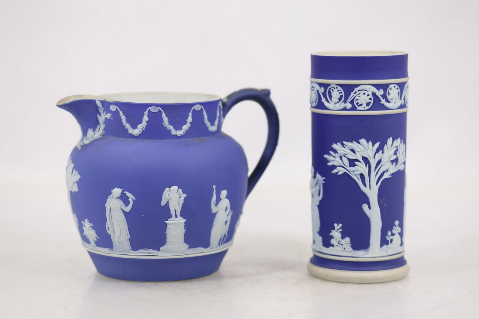 Blue Jasperware Pitcher and Cylindrical Vase: A pair of blue jasperware vessels includes a rounded pitcher and a cylindrical vase. Both have matte cobalt blue grounds with applied white neoclassical relief decoration depicting draped figures tree