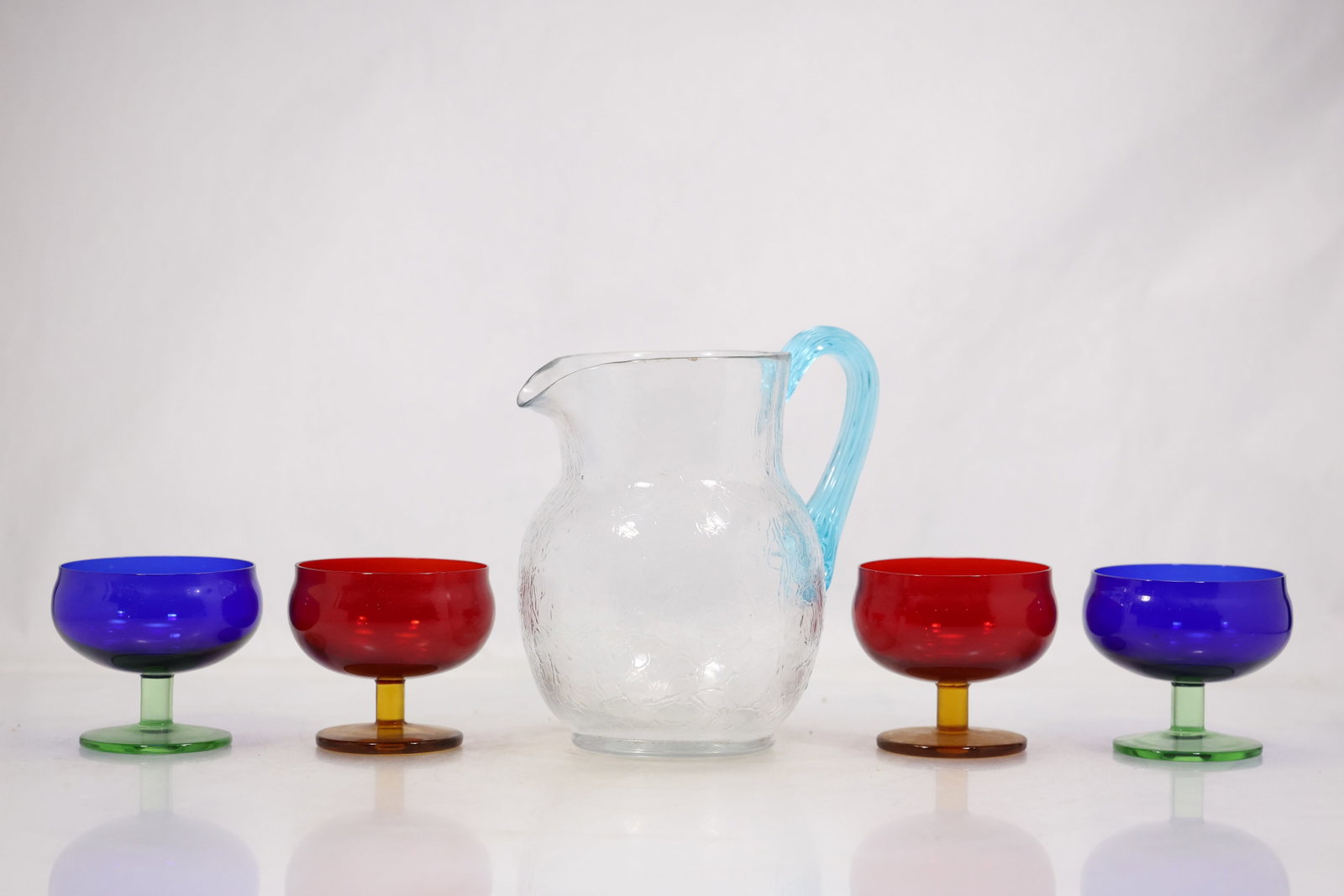 Multicolor Stemmed Glasses and Clear Pitcher Set: A beverage set includes a clear handled pitcher and stemmed glasses in multiple colors. The repeated bowl and stem forms with a coordinating serving pitcher identify the pieces as an assembled drinkwa