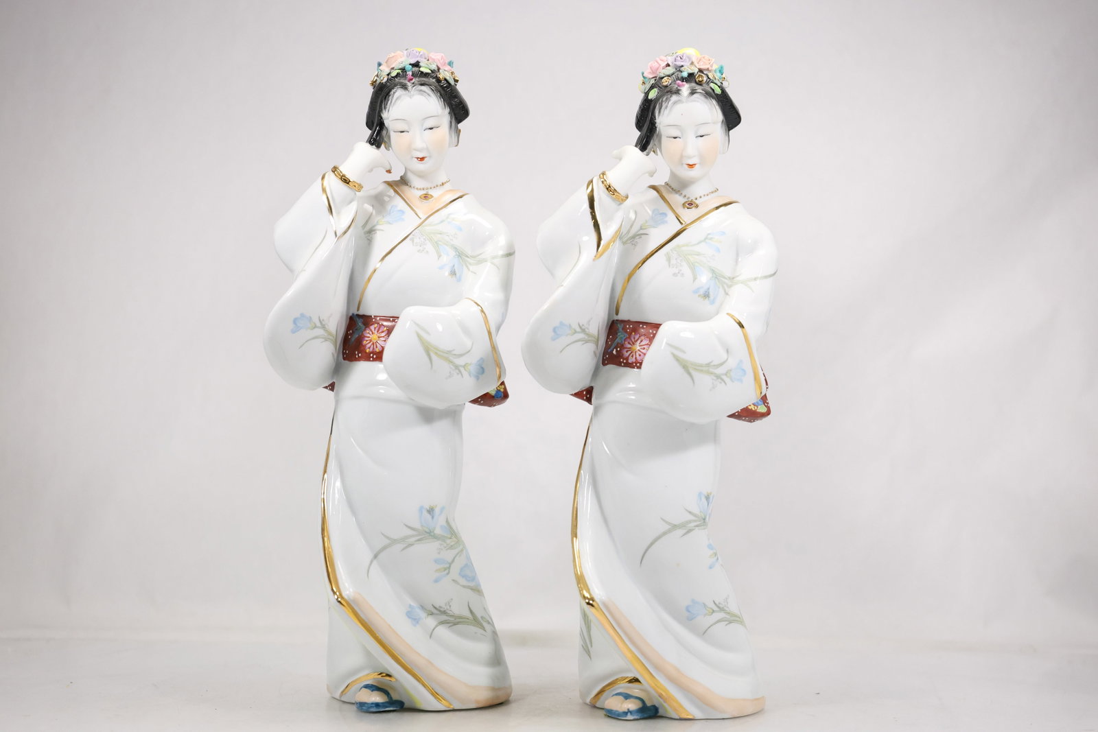 Pair Japanese Ceramic Woman Figurines: A pair of glazed ceramic figurines depicts Japanese women in traditional dress with painted facial details and colored robes. A red and gold foil label is visible but is not legible enough in the phot