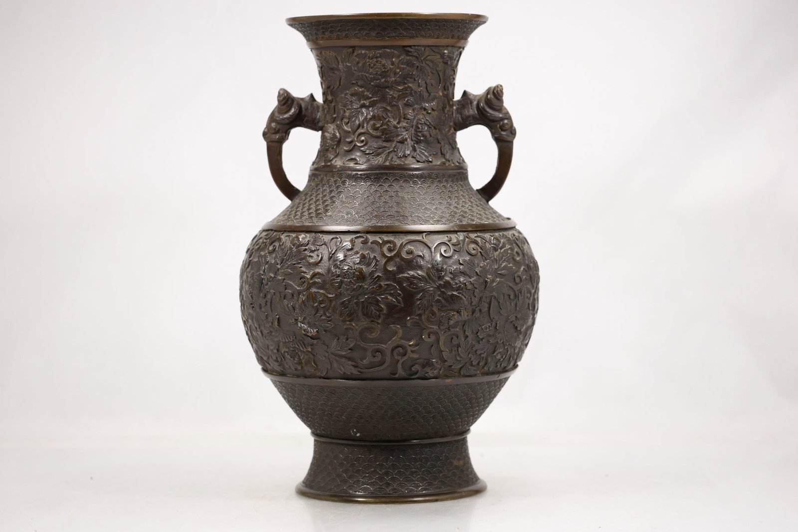 Etched Brass Double Handle Urn Vase: A brass urn form vase has paired handles a flared foot and etched ornamental bands across the body. The metal construction symmetrical silhouette and engraved surface identify it as decorative brasswa