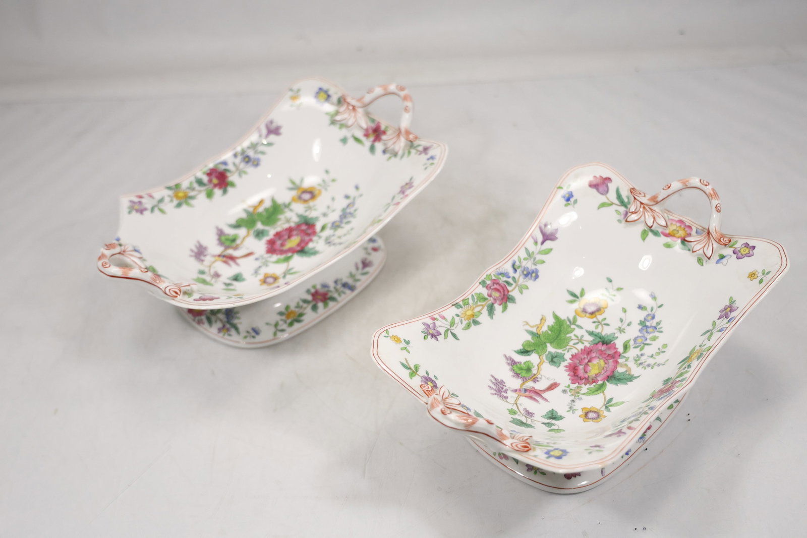 Pair Floral Porcelain Footed Serving Dishes: A matched pair of porcelain serving dishes has shaped bowls raised on small feet with multicolor floral decoration. The repeated form and coordinated botanical pattern identify them as tableware and n