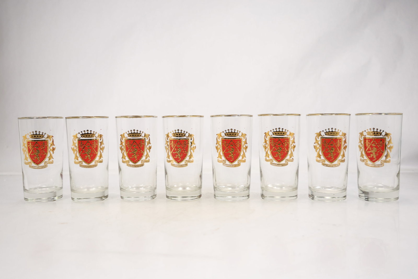 Set of Eight Red Shield Motif Glass Tumblers: A set of eight clear glass tumblers carries a repeated red shield shaped emblem on each cylindrical body. The matched form and coordinated heraldic motif identify them as a barware set and no maker