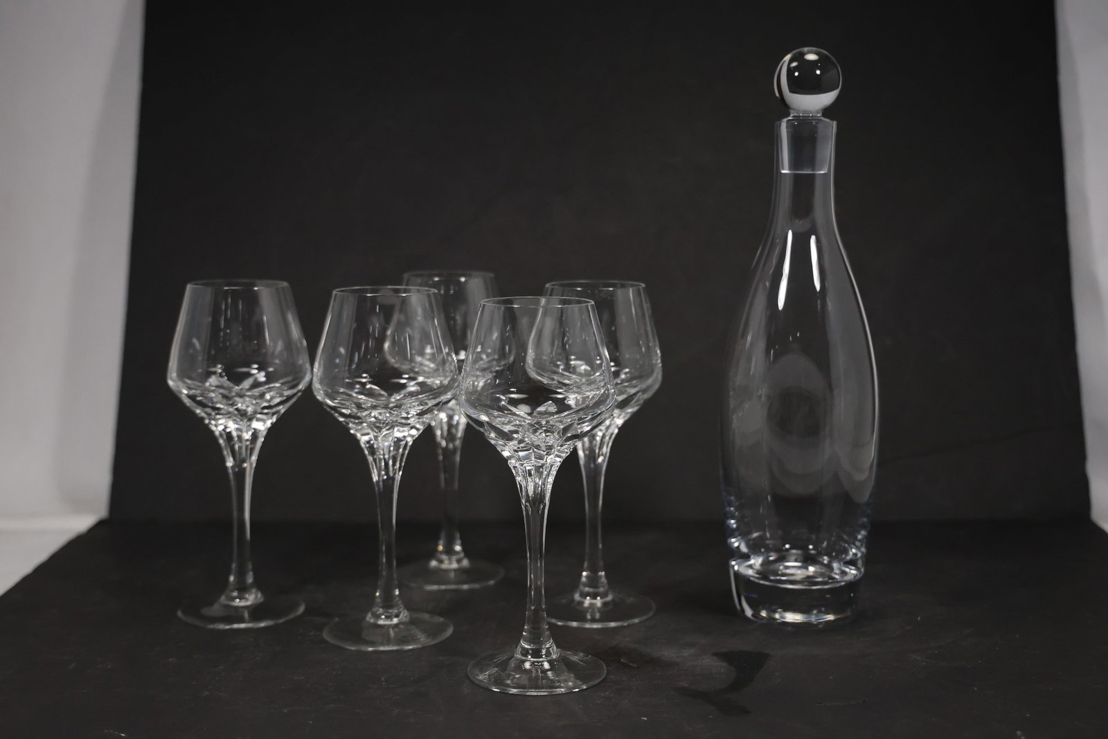 Signed Clear Glass Decanter and Four Stemmed Cups: A five piece clear glass beverage set includes one tall stoppered decanter and four matching stemmed cups. The coordinated tapered forms and an etched cursive signature visible on the base support a