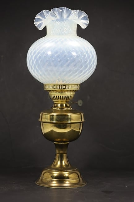 White Opaline Glass Oil Lamp with Brass Fittings: A traditional oil lamp has a white opaline glass font with molded ribbing and a ruffled upper edge joined to brass tone burner and base fittings. The reservoir burner mount and period lamp form identi