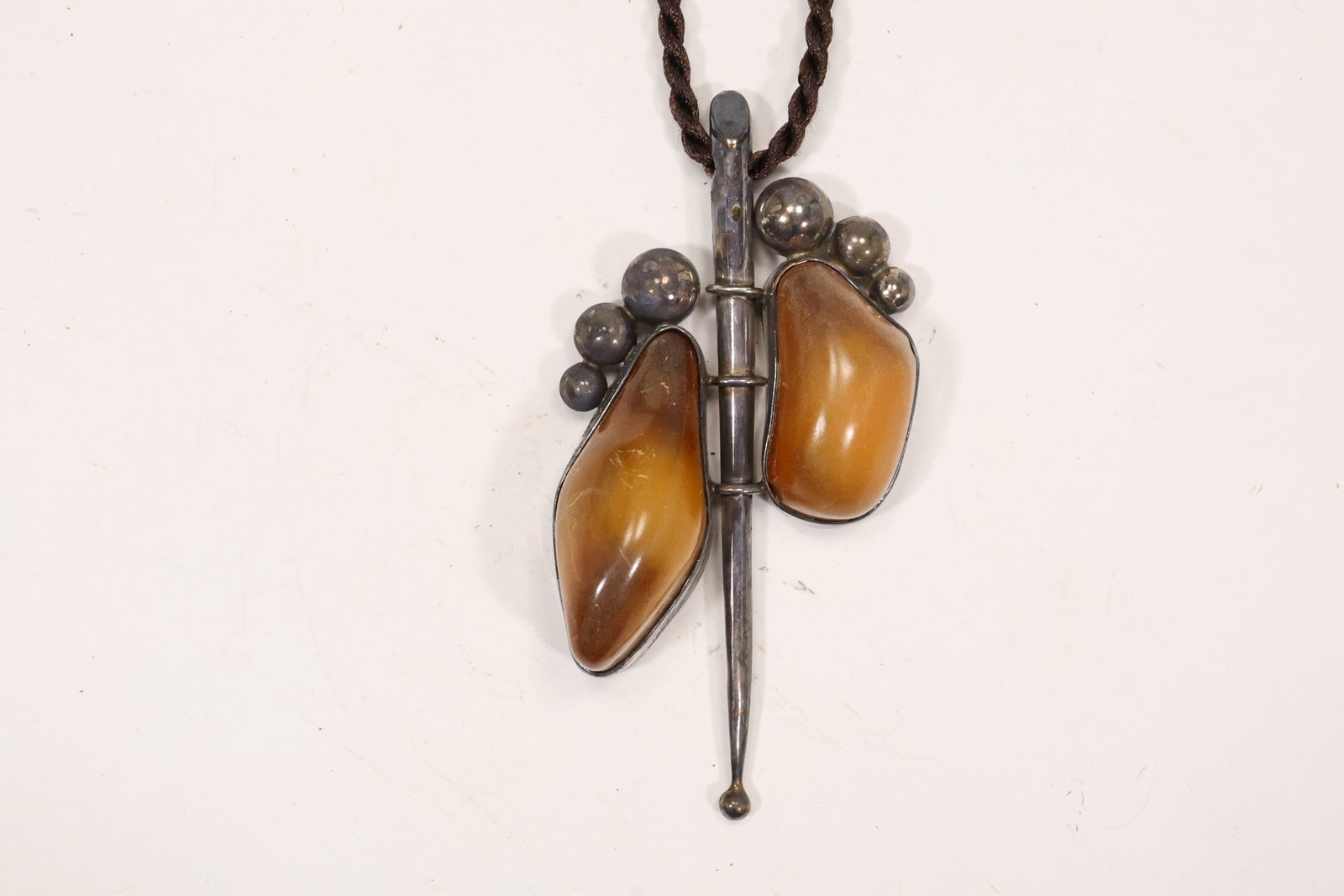 Sterling Silver Modernist Double Amber Pendant Necklace: A .925 sterling silver necklace has a large sculptural pendant set with two amber cabochons on a dark cord or rope. The asymmetrical openwork form and warm stones support a mid century modernist