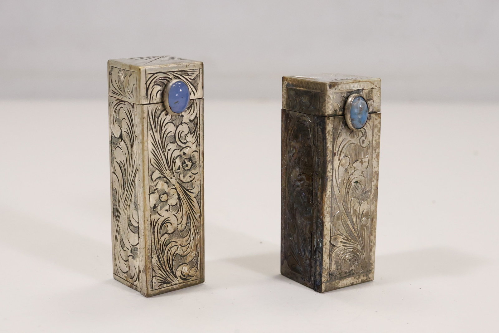 Pair Italian 800 Silver Gemstone Lipstick Compacts: A pair of 800 silver lipstick compacts has engraved rectangular cases set with a color change style cabochon and a turquoise cabochon. The hinged vanity accessory forms show scrolling surface decorati