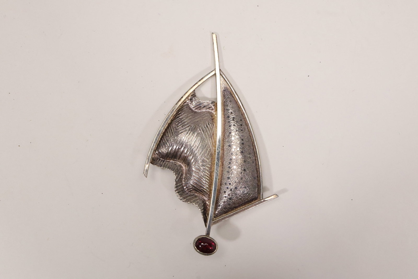 Sterling Silver Sailboat Pin with Red Stone: A sterling silver pin is modeled as a stylized sailboat with openwork sails curved hull and a small red stone accent. The long vertical profile and nautical subject are visible in the photographs. The