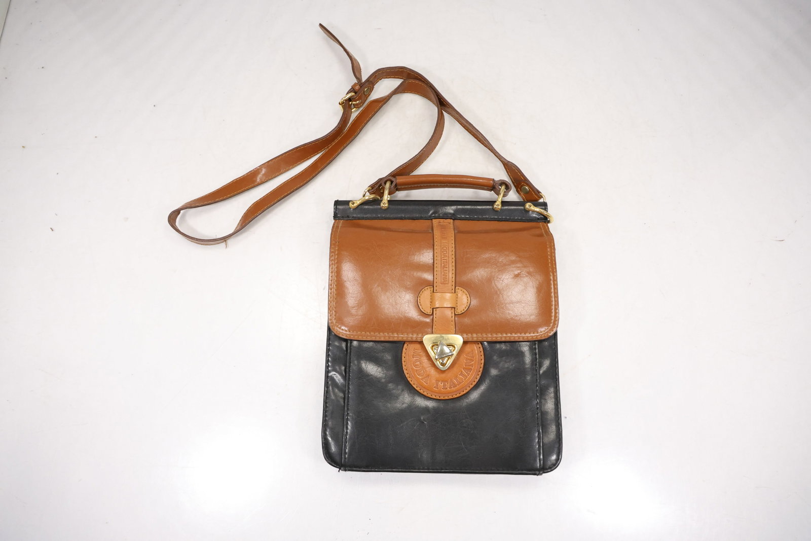 Black and Tan Leather Crest Crossbody Satchel: A small crossbody satchel combines a black body with tan leather flap trim shoulder strap and a triangular crest form clasp. No legible maker mark is confirmed in the photographs. The handbag construc