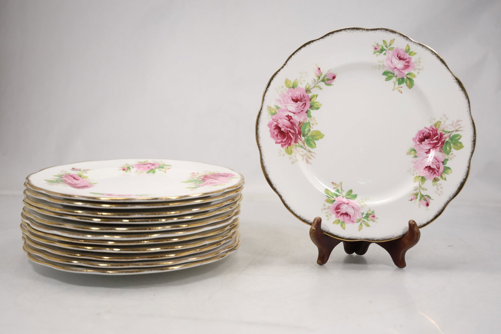 Set of Twelve Pink Rose Floral Porcelain Plates: A set of twelve porcelain plates has scalloped rims with dark line borders and sprays of pink roses with green foliage. The coordinated tableware set appears unmarked or lacks a legible maker mark in 