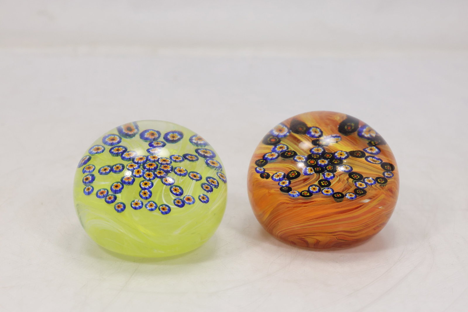 Two Colorful Millefiori Art Glass Paperweights: Pair of small domed art glass paperweights with bright yellow orange green and blue millefiori style floral inclusions. Each compact weight displays clustered cane patterns beneath clear glass. The fo