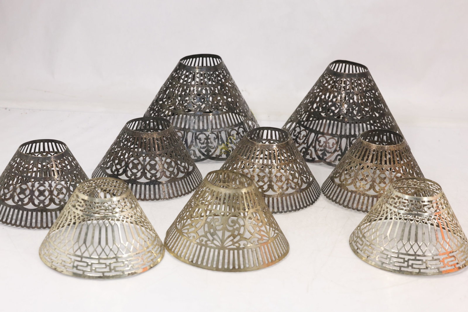 Nine Perforated Metal Lamp Shades: Group of nine matching flared metal lamp shades with perforated openwork patterns and scalloped lower edges. The silvery surfaces and repeated pierced motifs create a coordinated lighting component