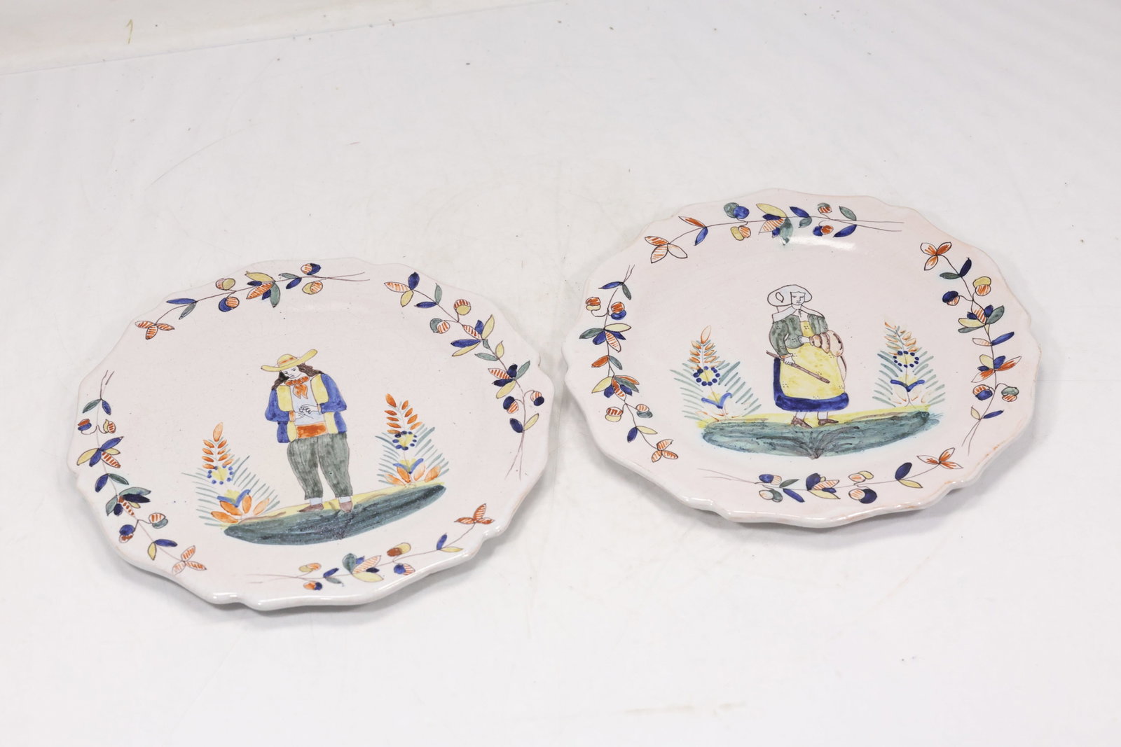 Pair Hand Painted Folk Art Porcelain Plates: Pair of porcelain plates decorated with colorful hand painted figures buildings and floral border motifs. The coordinated circular scenes have a European folk art character. The modeling relates to