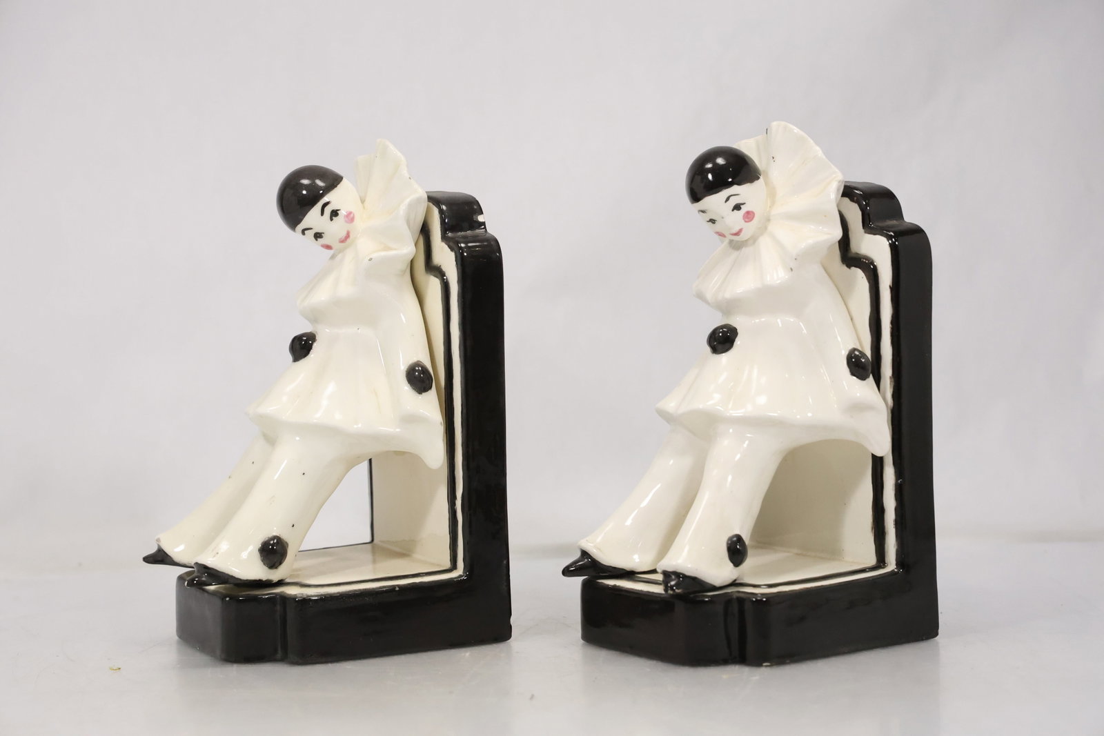 Pair Art Deco Style Clown Bookends: Pair of ceramic or composition bookends modeled as seated clowns in black white and cream. Their geometric costumes and stylized profiles evoke Art Deco theatrical design. The subject fits twentieth