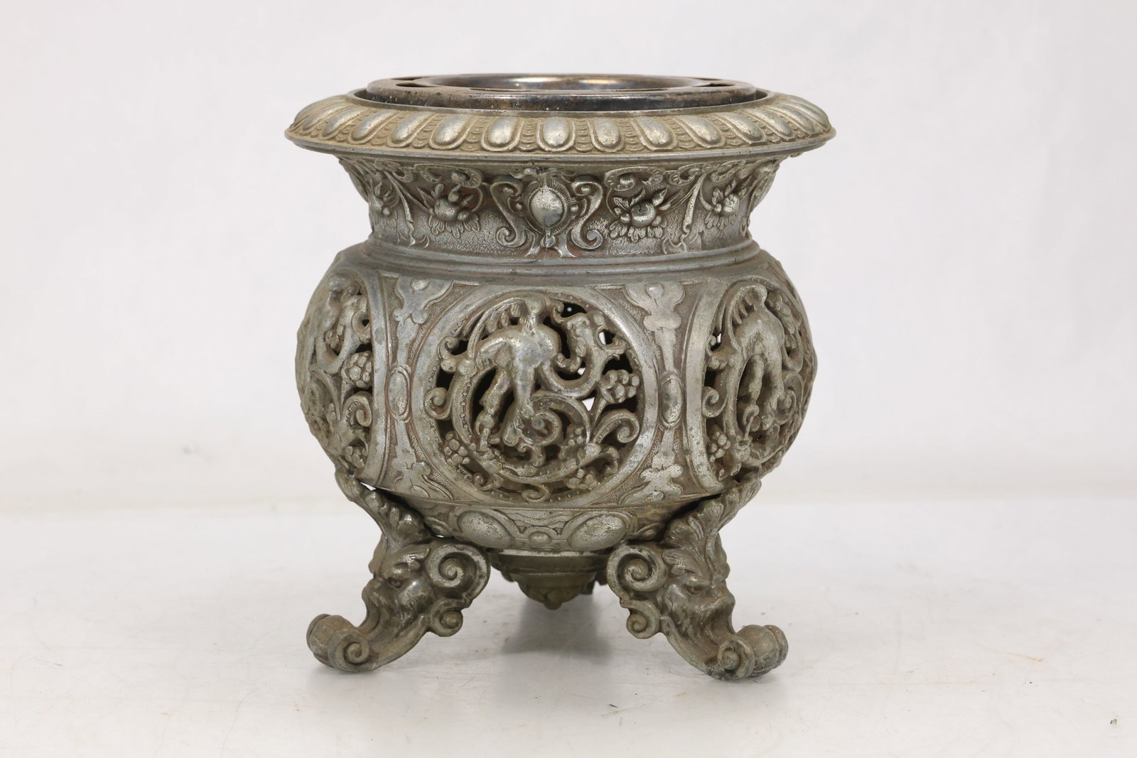 Ornate Silver Tone Repousse Urn: Decorative silver tone urn with pierced scrolling panels floral repousse ornament and four elaborate feet. The rounded body and flared rim create a richly detailed revival style vessel. The design