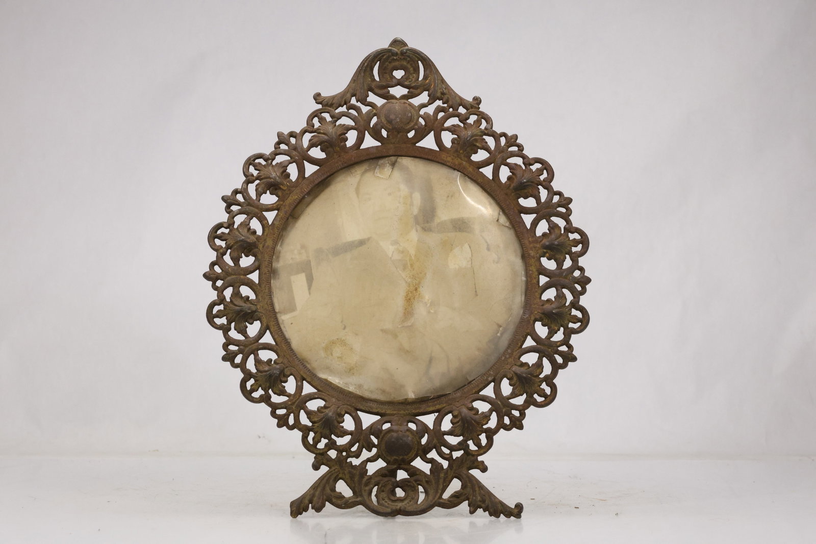 Ornate Cast Metal Oval Picture Frame: Oval tabletop picture frame with pierced cast metal border floral scrollwork and prominent crest. The bronze tone surface and easel form suit a portrait or miniature. The design relates to decorative