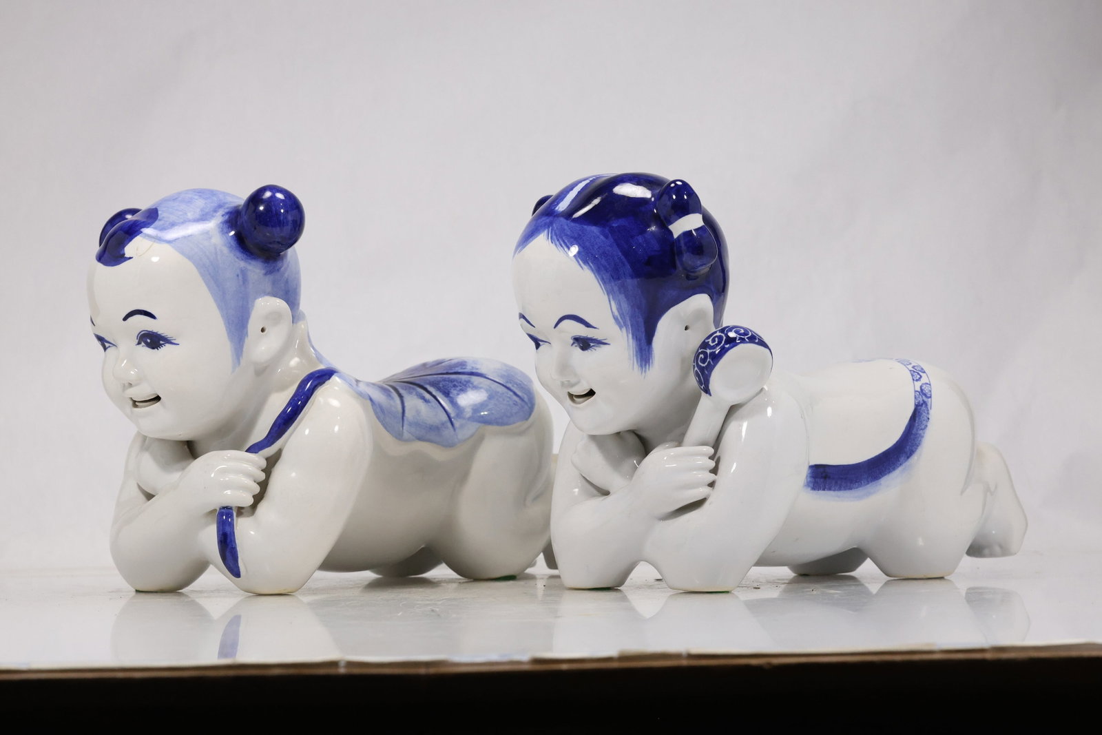 Pair Chinese Blue and White Porcelain Child Figures: Pair of glazed porcelain child figures modeled reclining on their stomachs with blue hair clothing accents and playful expressions. The coordinated figures follow Chinese decorative ceramic figure