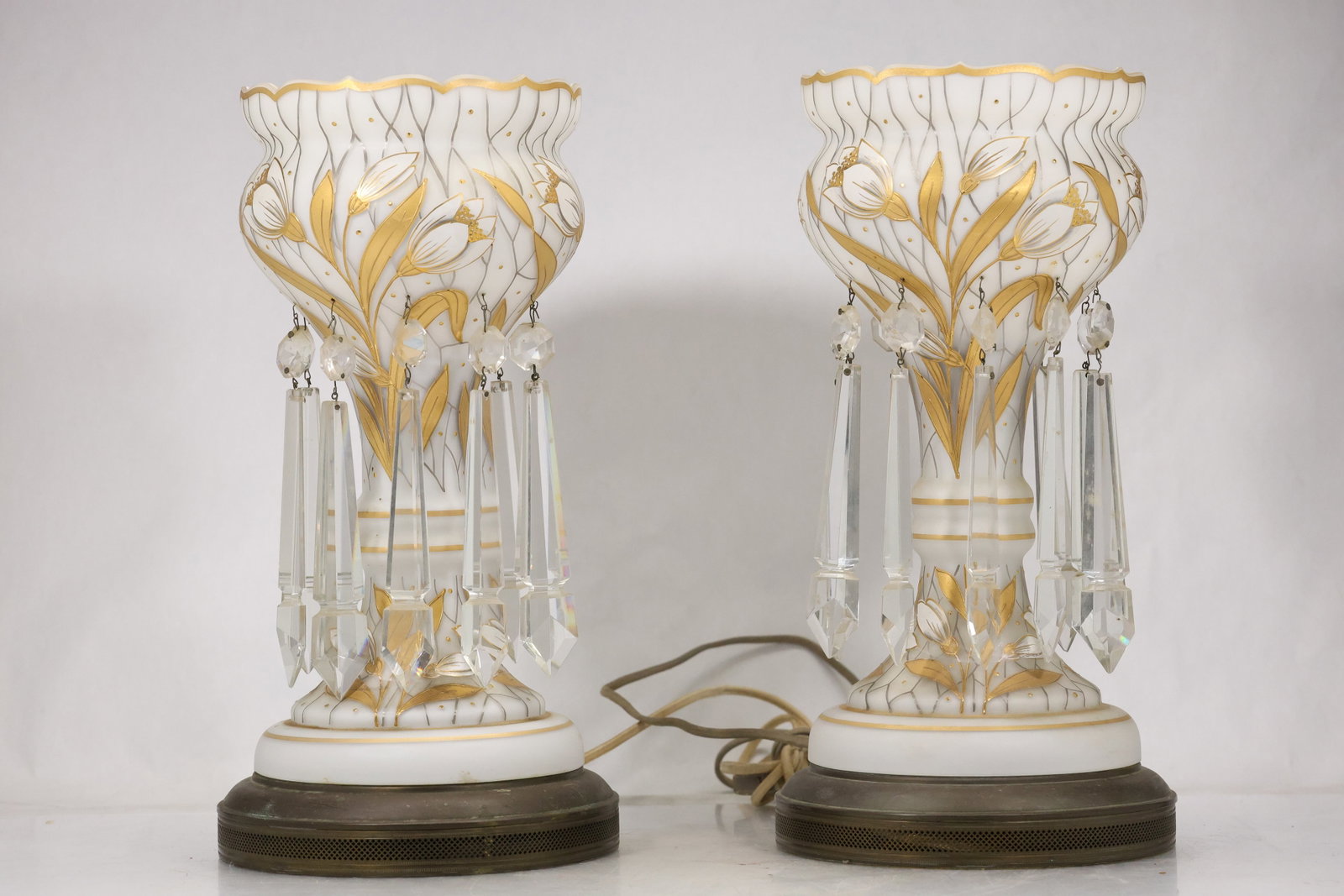 Pair Gilt Floral Crystal Table Lamps: Pair of tall table lamps with clear faceted crystal columns white porcelain collars hand painted gilt leaf decoration and round dark wood bases. Each lamp is wired with a cord and the open upper socke