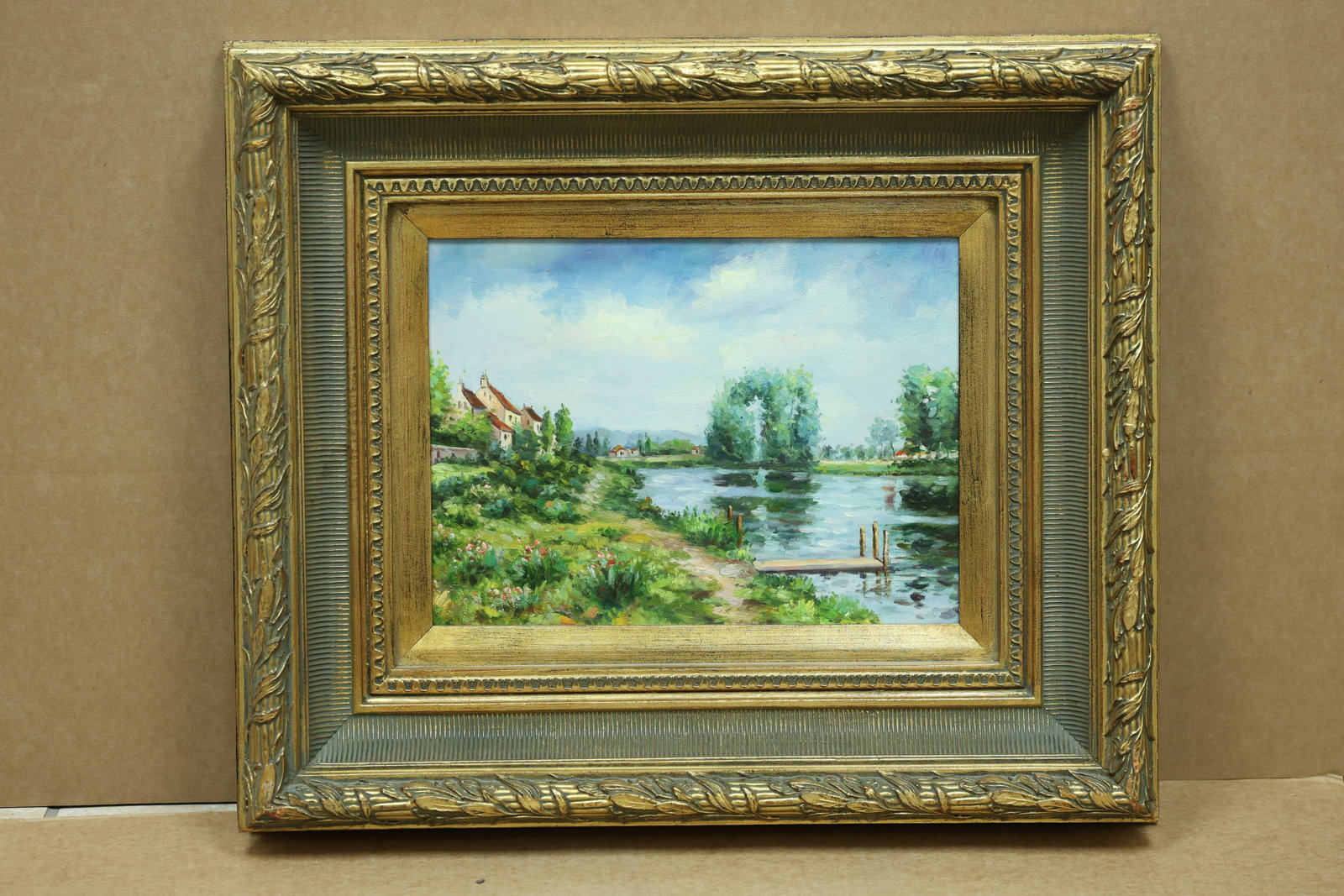 European River Cottages and Dock Landscape Painting: Framed landscape painting depicting cottages and a dock beside a river in an ornate frame. No readable artist signature is visible in the reviewed photographs. The composed view relates to American an
