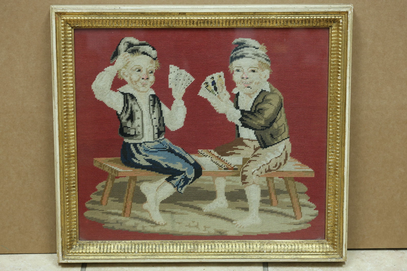 Two Boys Playing Cards Needlepoint on Red Ground: Framed needlepoint depicting two boys playing cards against a red ground. No readable maker or workshop mark is visible in the reviewed photographs. The stitched image relates to Arts and Crafts needl