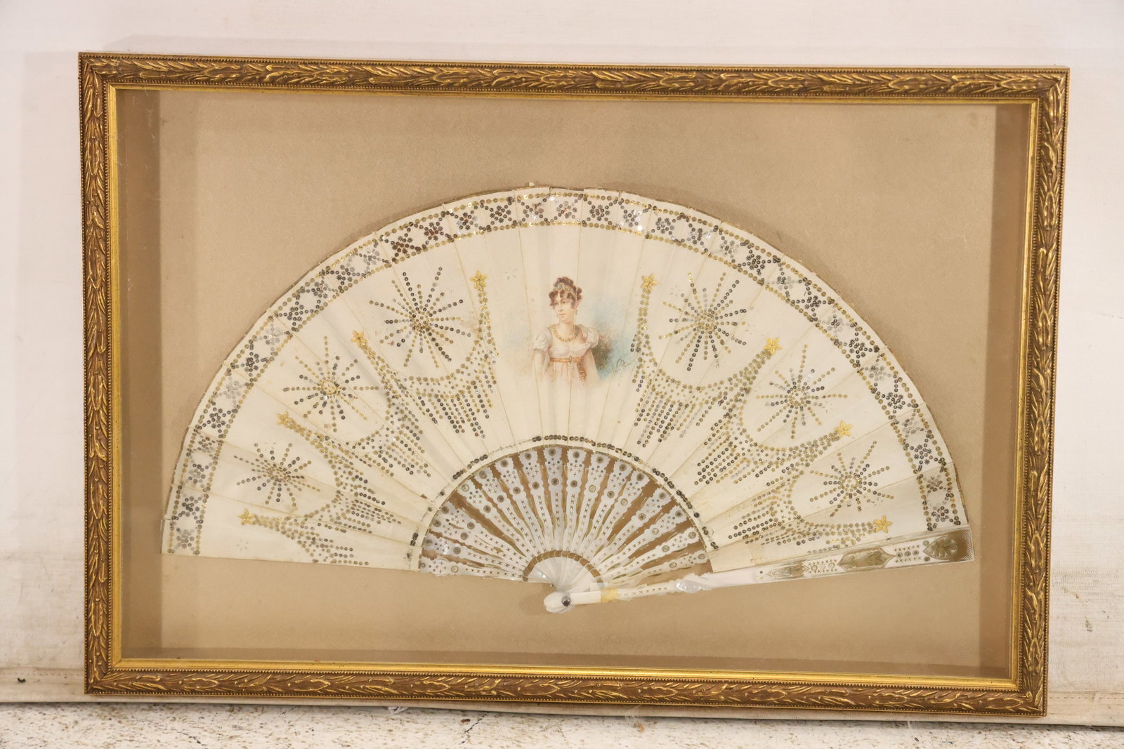 Antique Hand Painted Mother of Pearl Folding Fan in Frame: Antique folding fan mounted in a deep decorative frame. The fan has pierced mother of pearl tone sticks and a hand painted leaf with a central female portrait floral sprays and radiating gilt decorati
