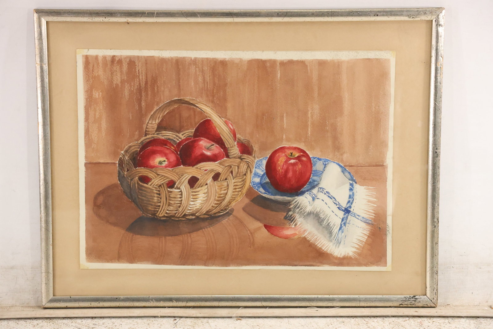 Signed D J Manning Apple a Day Watercolor Still Life: Original watercolor titled Apple a Day by D J Manning. A wicker basket filled with red apples rests beside a single apple on a blue and white plate and a folded embroidered cloth. The work is signed a
