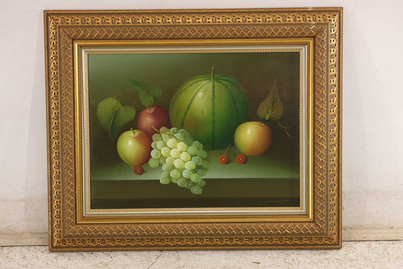 Signed Gianni Fruit Still Life Oil Painting: Oil on canvas depicting watermelon, green grapes, apples, pears, and cherries arranged on a ledge. Signed “Gianni” lower right. Presented in an ornate gold-toned frame. Artwork measures 13 x