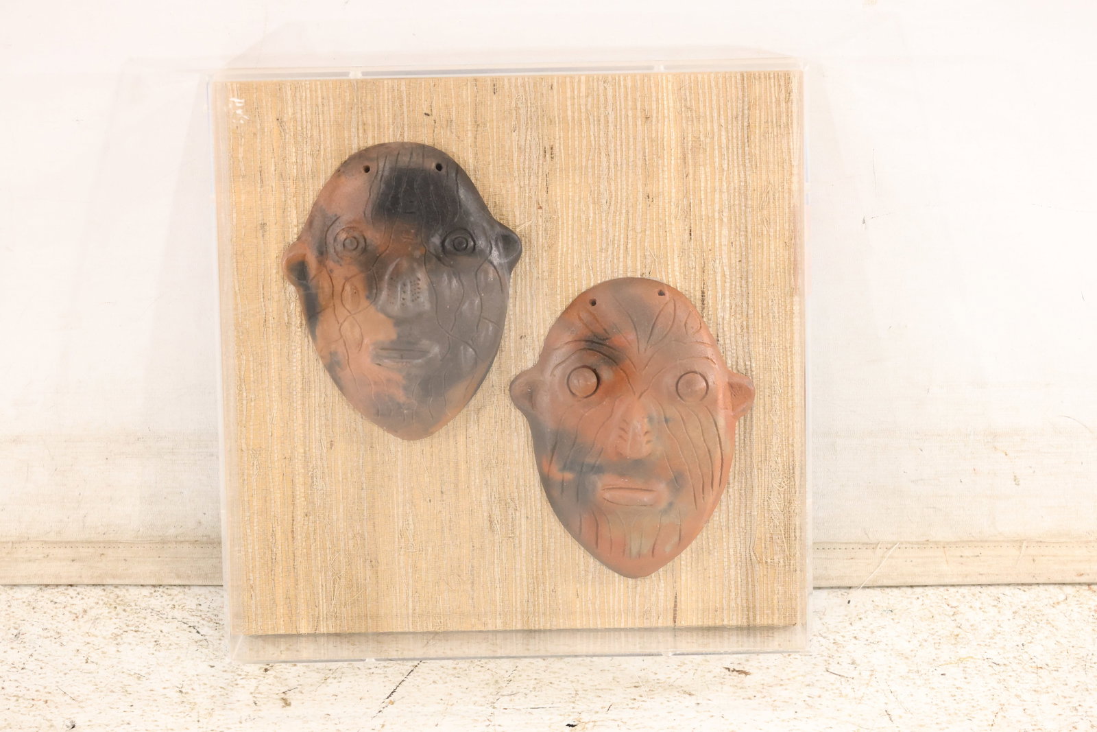 Two Terra Cotta Face Masks in Shadow Box: Two terra cotta face masks or amulets in a Pre-Columbian style, with incised details and pierced suspension holes. Mounted on fabric backing in a clear acrylic display case. Display measures 13.5 x 13