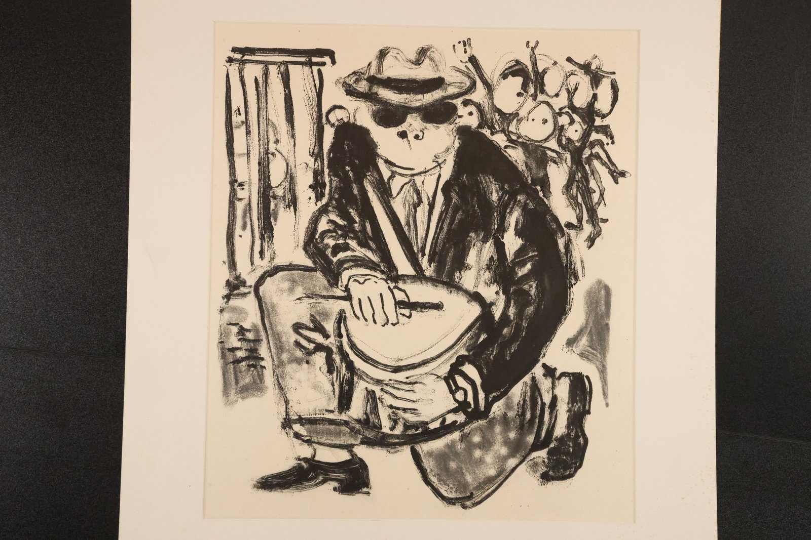 Black-and-White Lithograph of Drummer in Hat and Glasses: Black-and-white lithograph depicting a seated drummer wearing a hat and dark glasses, with loosely drawn figures and foliage behind. Presented with a light-colored mat. Size: Outside mat: 26 x 22 inch