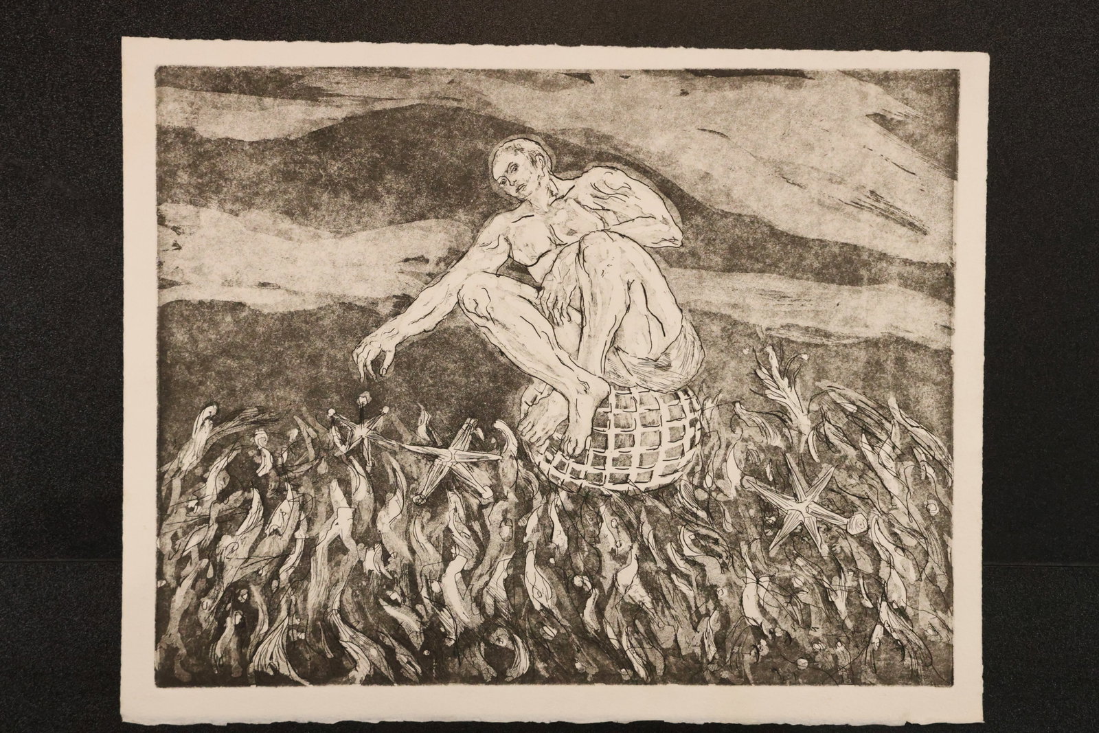 Figure on a Basket Black-and-White Print: Black-and-white print depicting a seated figure on a woven basket, reaching toward a field of flame-like marks and scattered jack-shaped forms beneath a dark sky. Size: 12 x 15 inches. Shelf ID: mc2 *