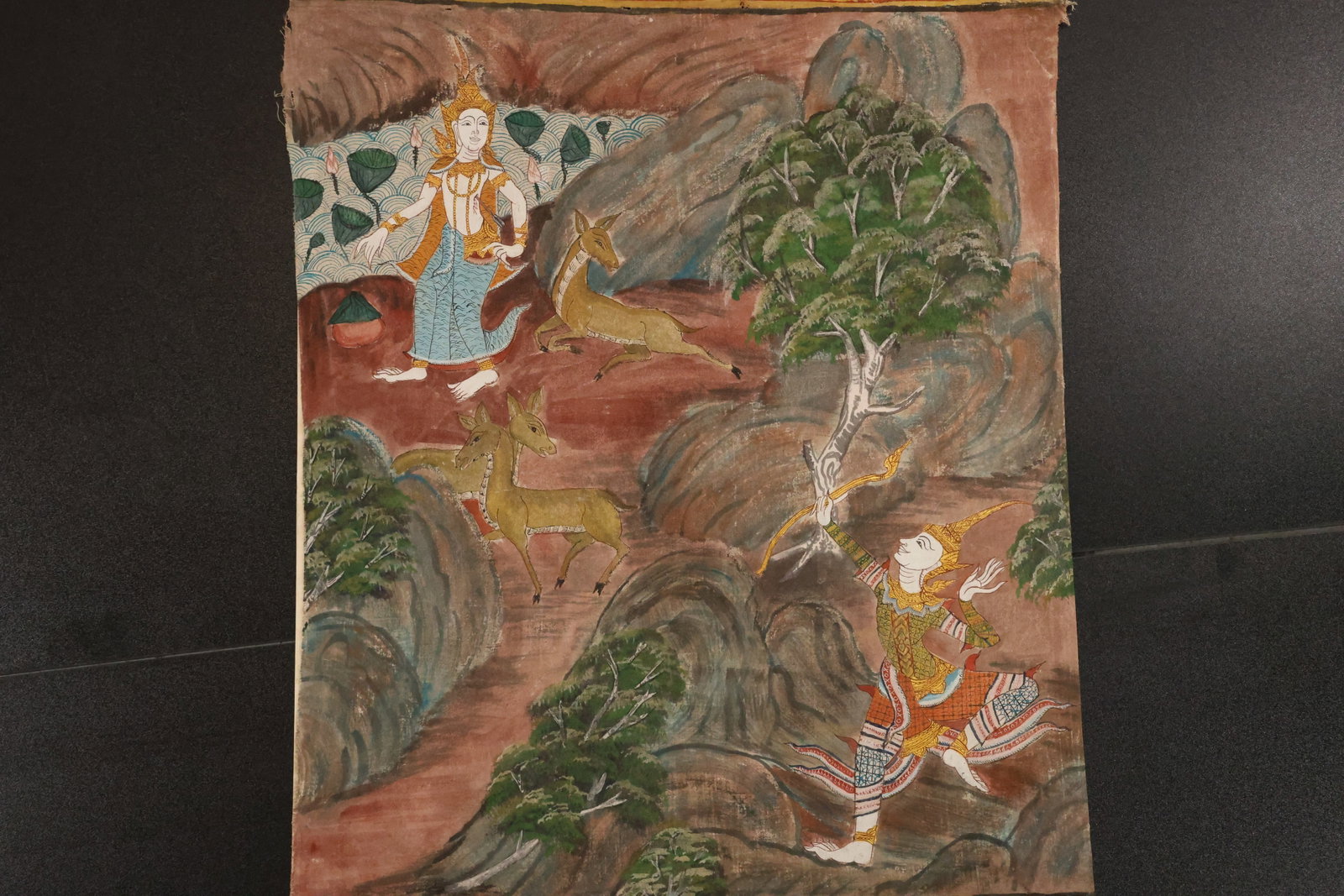 Traditional Figural Landscape Painting on Canvas: Painting on canvas wrapped over a board, depicting figures in traditional dress among rocky terrain, trees, and a pale arched structure. Rendered in earthy red, green, blue, and ochre tones. Size: 22 