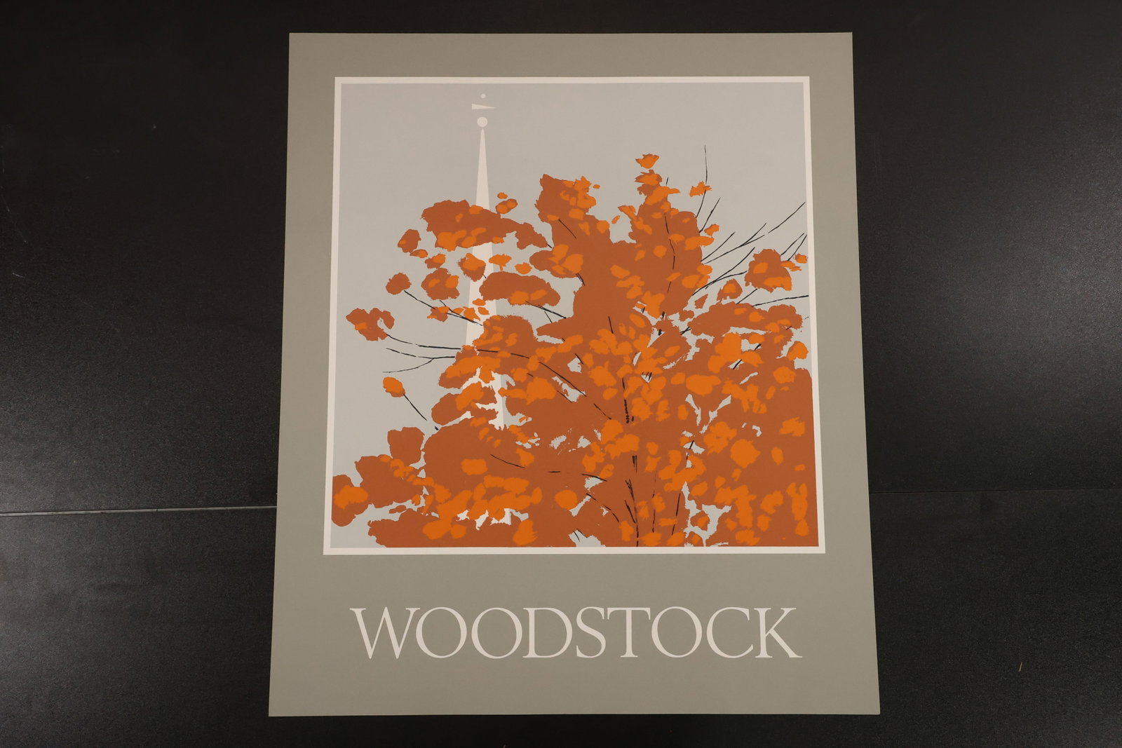 R. Angeloch Woodstock Print with Gray Lettering Panel: A color print by R. Angeloch featuring orange foliage and a pale steeple against gray. WOODSTOCK appears in light lettering on the darker panel beneath the image. Size: 26 x 22 inches. Shelf ID: mc4 *