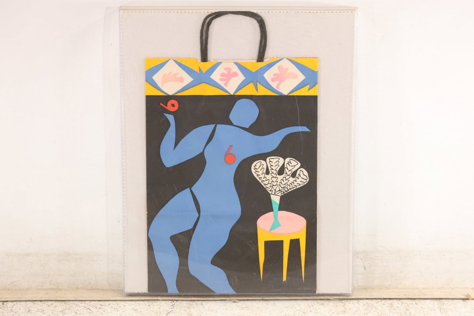 Bloomingdale’s Blue Figure Shopping Bag in Shadow Box: A decorative paper shopping bag identified in the accompanying notes as a Bloomingdale’s New Year 1982 design. A stylized blue figure appears beside a yellow stool and a vase of white foliage, benea