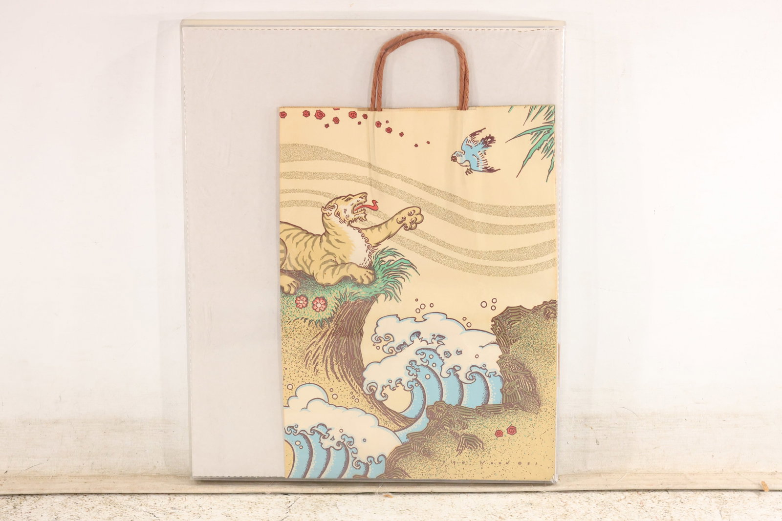 Page Wood Design Tiger Shopping Bag in Shadow Box: A decorative shopping bag with a tiger-and-bird design attributed to Page Wood for Bloomingdale’s. A tiger reaches toward a small flying bird above curling blue waves, rocky cliffs, and scattered bl
