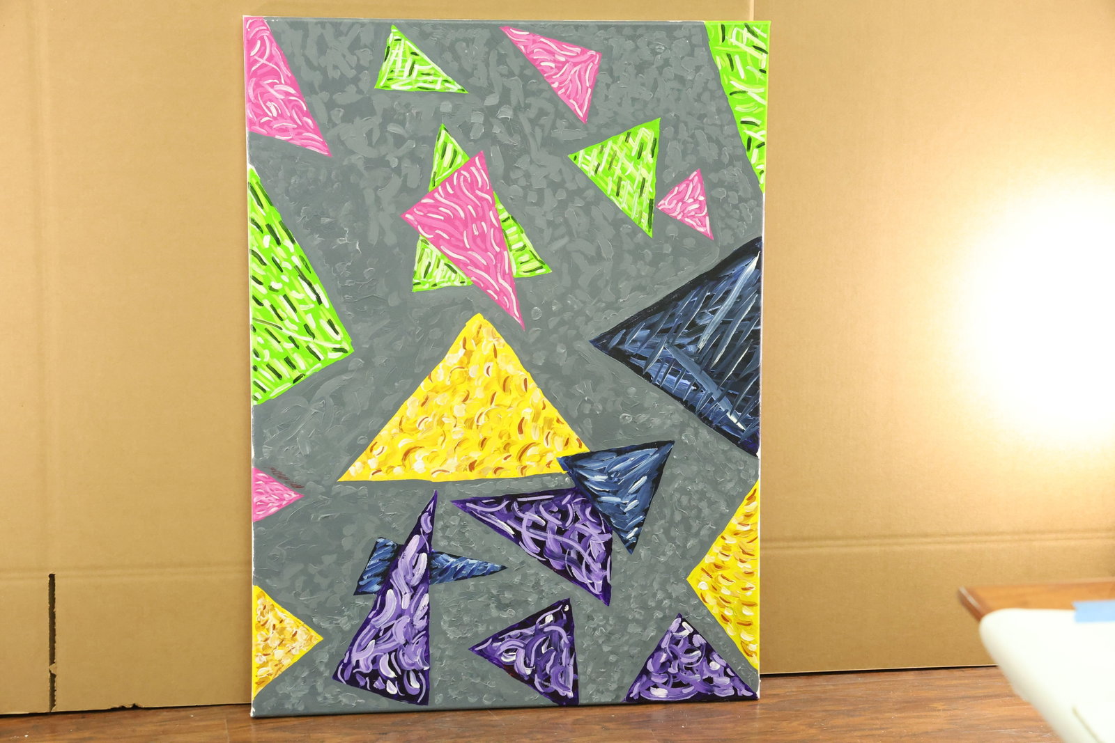 Geometric Abstract Triangle Painting on Gray Ground: Vertical canvas painting with scattered triangles in lime green, pink, yellow, blue and purple against a patterned gray ground. Light colored strokes and ridges fill the triangular shapes, with severa