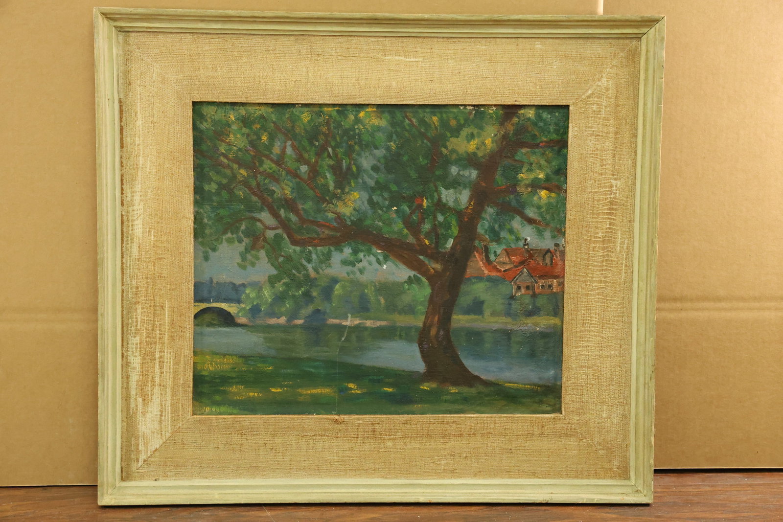 Framed Riverside Landscape Painting with Tree and Bridge: Landscape painting centered on a spreading leafy tree beside water. An arched bridge appears at left, with red roofed buildings beyond the river at right. Green foliage, brown branches and blue water 