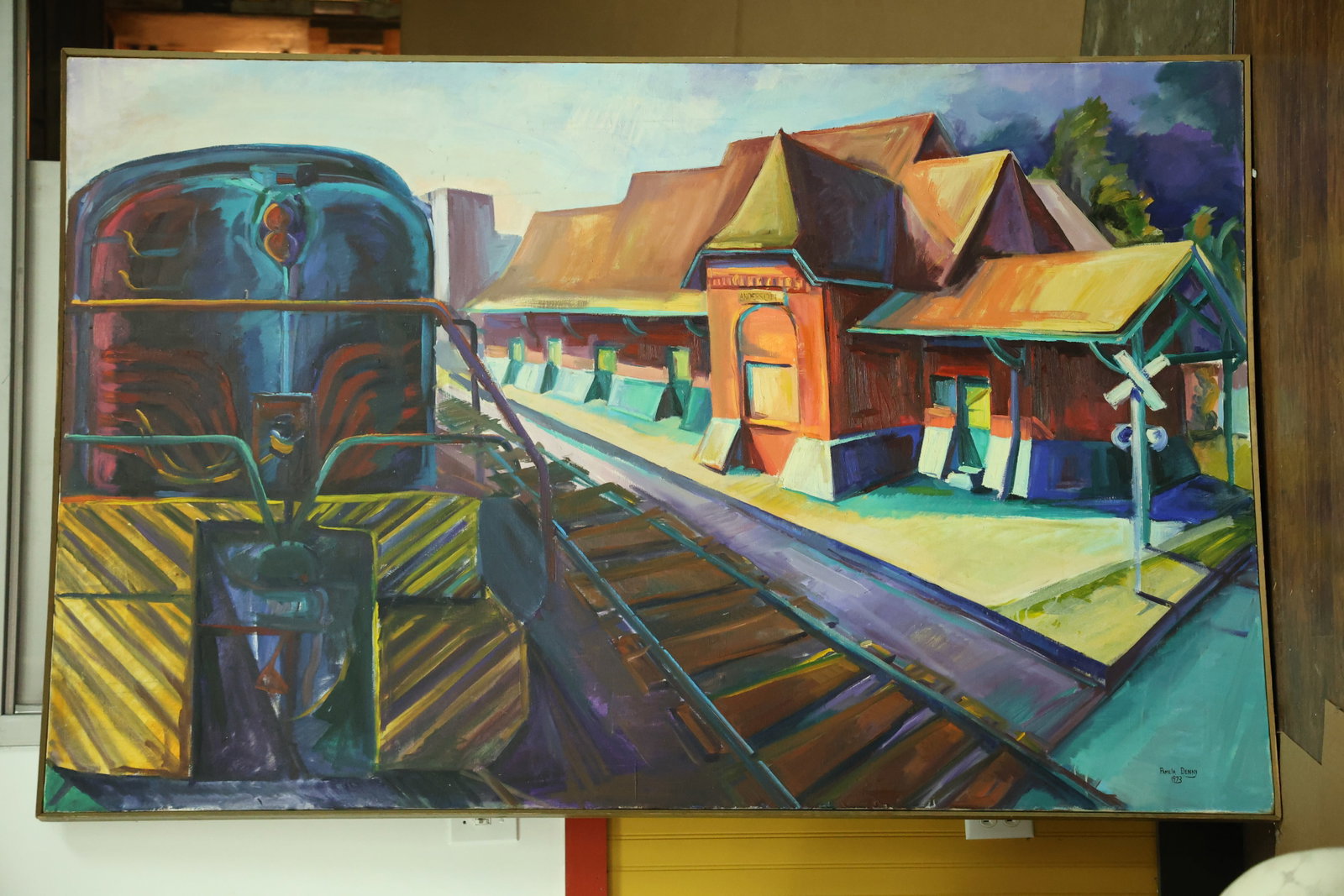 Railroad Locomotive and Depot Painting Signed Pamela Denny 1973: Large horizontal painting on canvas showing a locomotive beside converging railway tracks and a red orange depot building. Yellow and black stripes mark the engine front. Blue and purple shadows, a cl