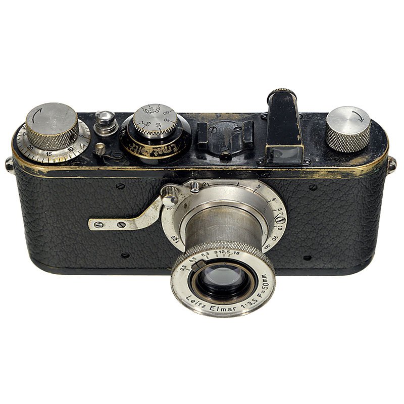 Leica I (A) with Elmar, 1927: Leica I (A) with Elmar, 1927 Leitz, Wetzlar. Early 4-digit serial no. 5042, Elmar 3,5/50 mm, cable release button, high-profile winding knob (8,5 mm), extensive vulcanite, dents in body. With original