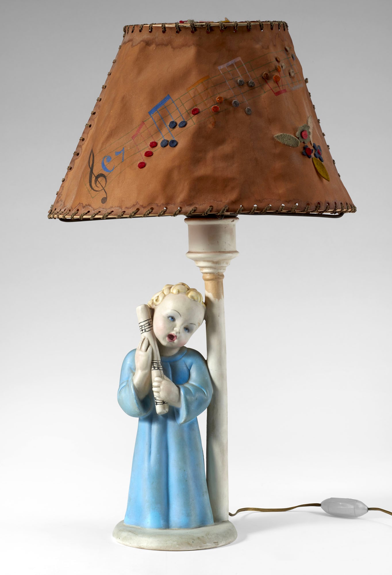 Lenci Ceramic table lamp decorated with a full-relief figure of a singing angel. Lampshade in: Lenci Ceramic table lamp decorated with a full-relief figure of a singing angel. Lampshade in parchment decorated with polychrome painted musical staves and felt appliqués. Turin, 1930s. (h 46 cm.) (