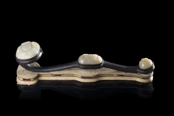 A ruyi sceptre with inset celadon jade plaques, carved: A ruyi sceptre with inset celadon jade plaques, carved with bats and auspicious Buddhist symbols, on a dark wood support, with original brocade base (some losses) China, 19th century (l. 34 cm.)