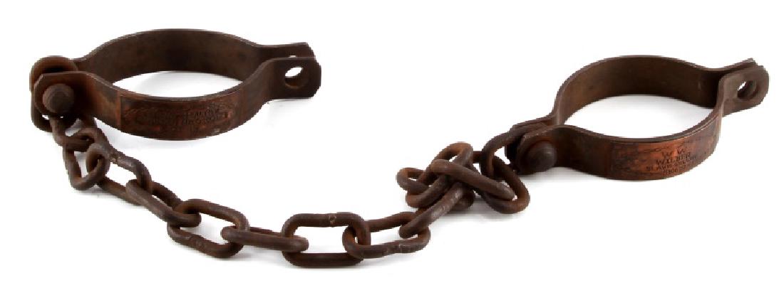 W W WILBER SLAVE SELLER CAST IRON CUFF SHACKLES: Cast iron slave shackles with copper plate engravings. One reads "W.W. WIlber Slave Seller 1806", the other reads "Strong Healthy African Negroes Lot 1A". 27" span. 3.5" x 2.75" cuff. Unauthenticated.