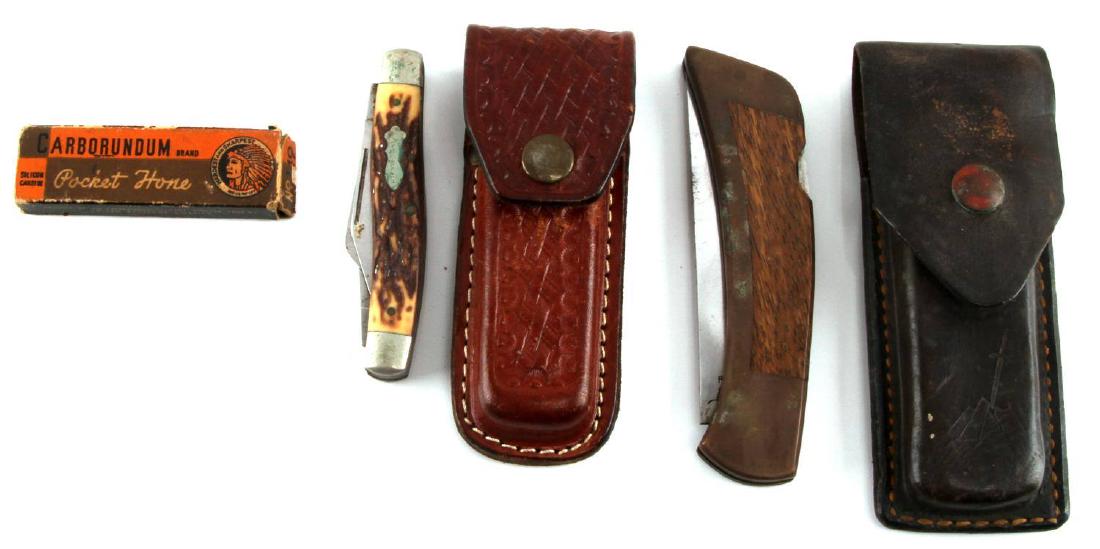 VINTAGE LEATHER CASE FOLDING POCKET KNIFE LOT: Two mid-sized folding pocket knives in leather pouches, as well as a honing stone in original box. Knives measure 4 and 5 inches in length closed, and the honing stone measures 3 inches in length. Ver