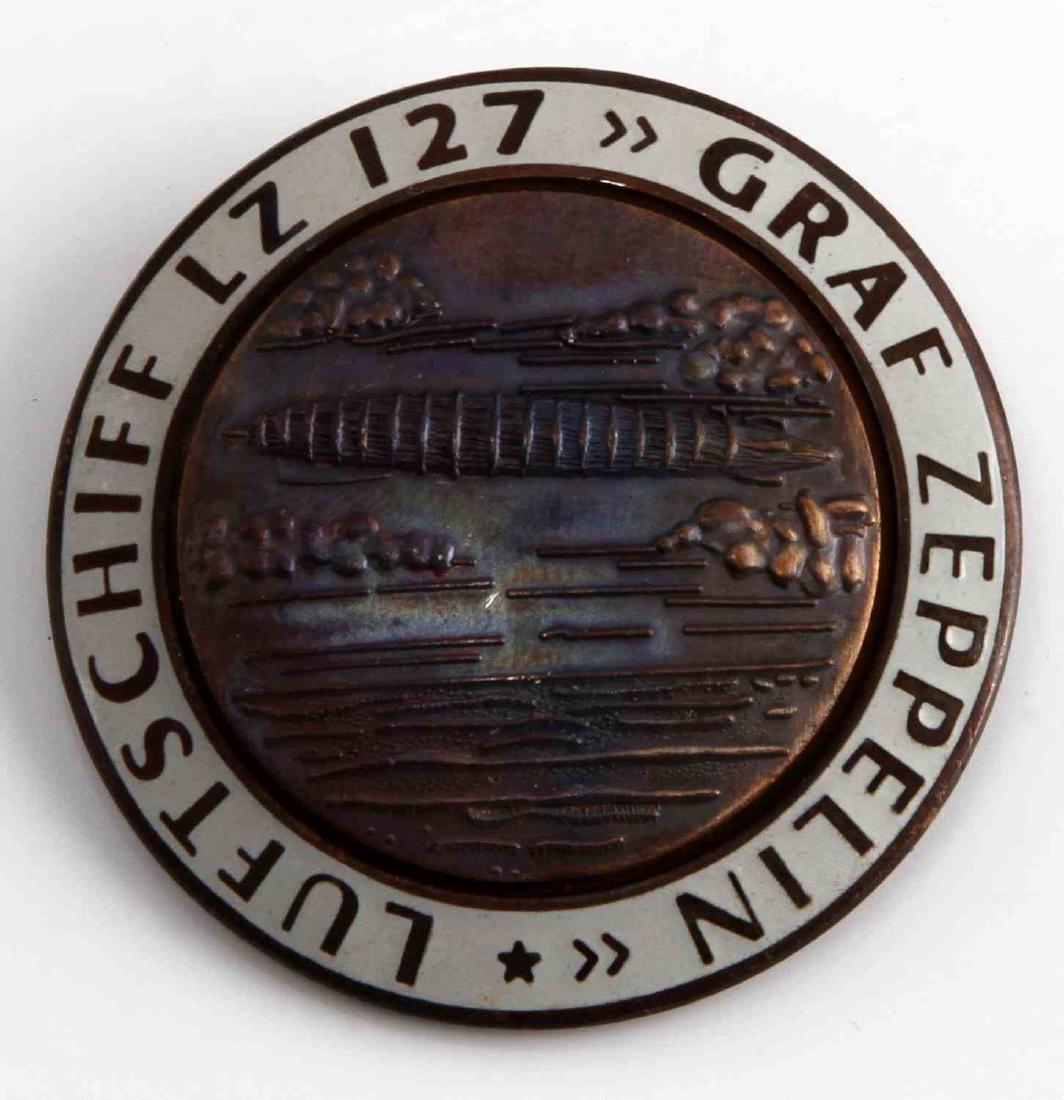 WWII GERMAN THIRD REICH GRAF ZEPPELIN TOUR BADGE: Graf Zeppelin LZ127 airship badge. Measures 1 3/4 inches in diameter. The front reads 'Graf Zeppelin Luftschiff LZ127' The front shows the airship flying over water. The reverse is maker marked 'FLL'.