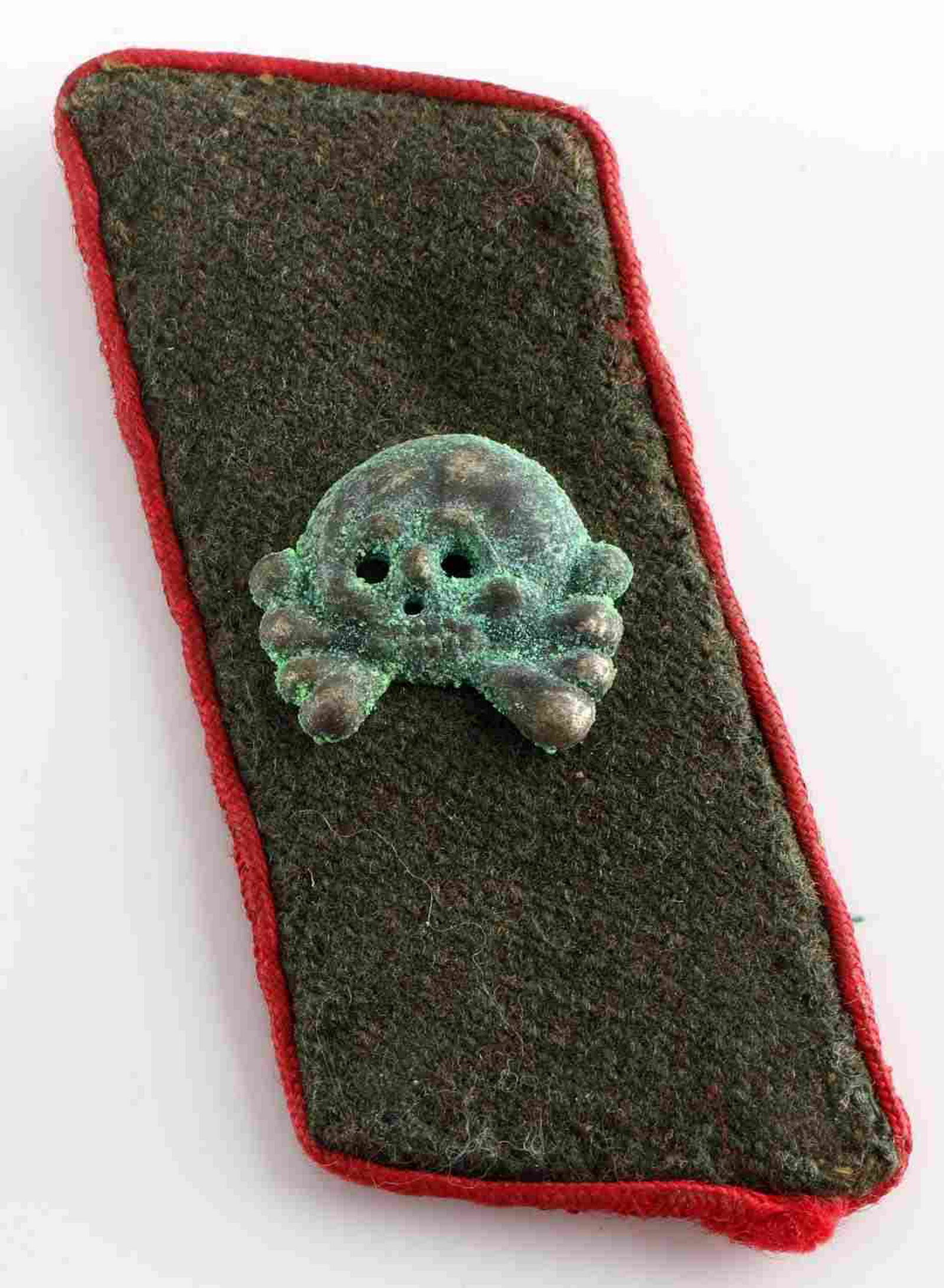 WWII GERMAN THIRD REICH ASSAULT GUNNER COLLAR TAB: Collar tab of a German enlisted man crewing an assault gun. Featurings a nickel jawless totenkopf on an OD green wool backing with artillery red piping around the edges. Reverse shows brown buckram re