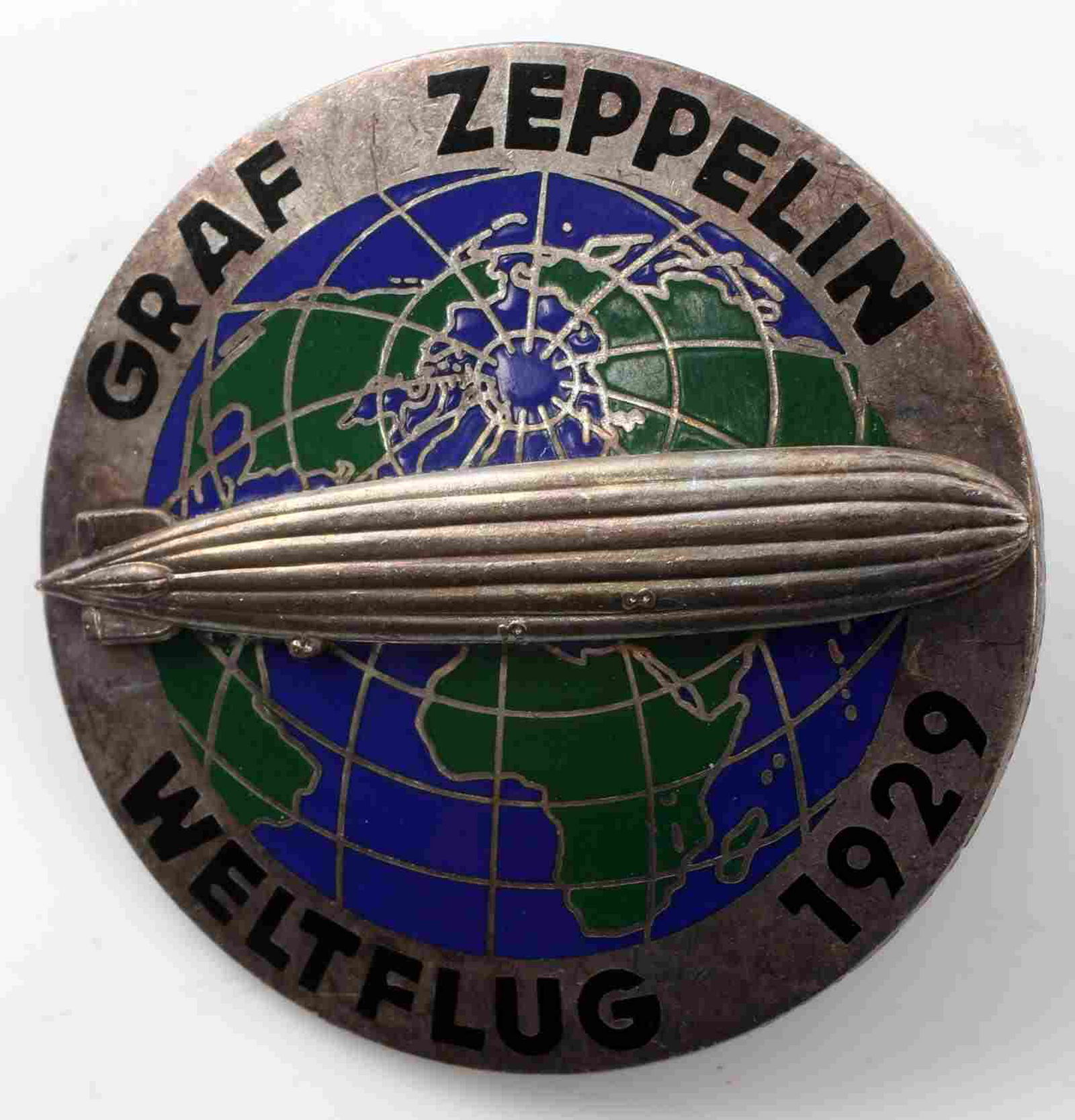 GERMAN GRAF ZEPPELIN WELTFLUG 1929 AIR SHIP BADGE: German WWII Graf Zeppelin Weltflug 1929 Air Ship Badge. Measures 1.75 inches in diameter. The front reads ‘Graf Zeppelin Weltflug 1929’. It shows the zeppelin flying over the world continents. The